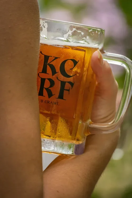 A person holding a glass mug of beer outdoors.
