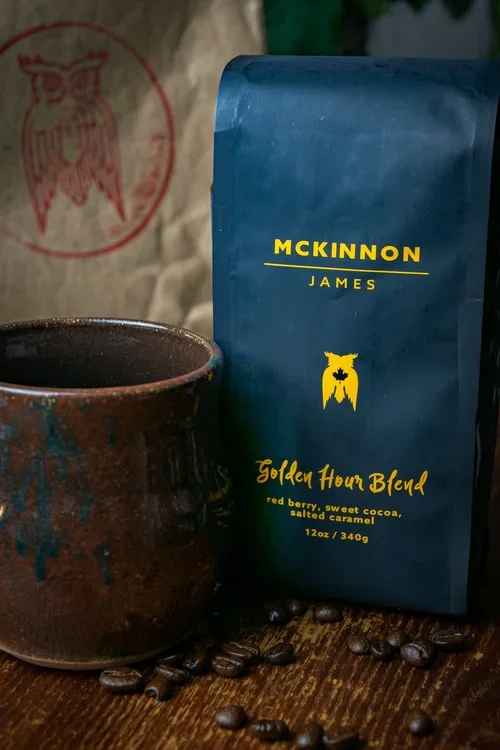 A bag of McKinnon James Golden Hour Blend coffee, with a dark ceramic mug and scattered coffee beans on a wooden surface.