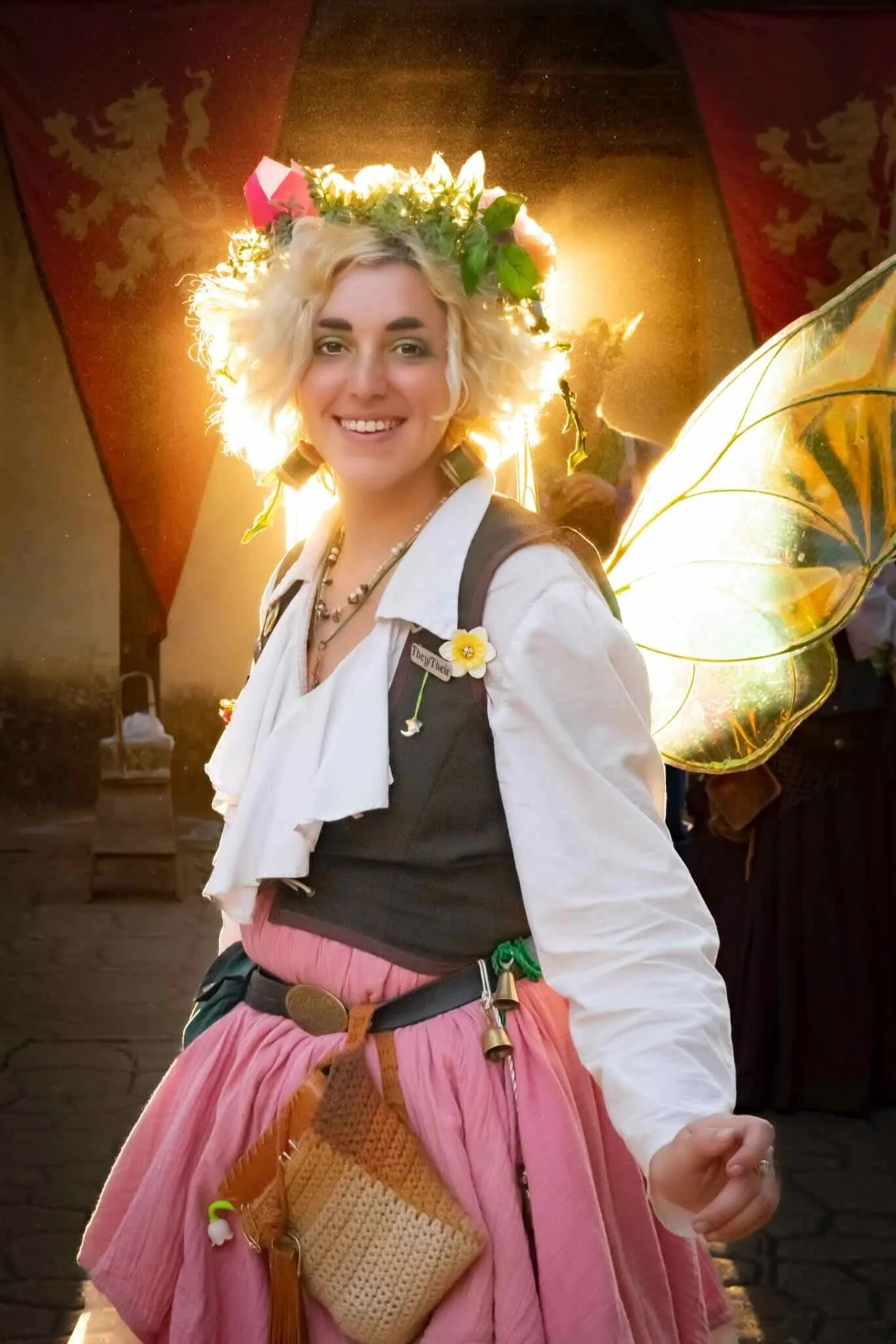 A smiling woman dressed as a fairy with butterfly wings, wearing a flower crown, standing indoors with warm lighting.