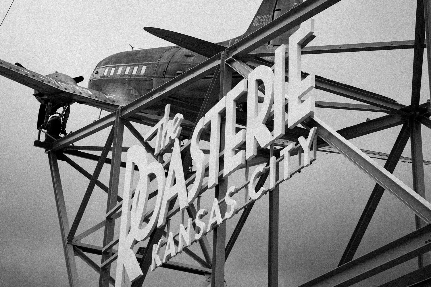 Black and white photo of the Roasterie Kansas City sign with a vintage airplane on a metal structure in the background.