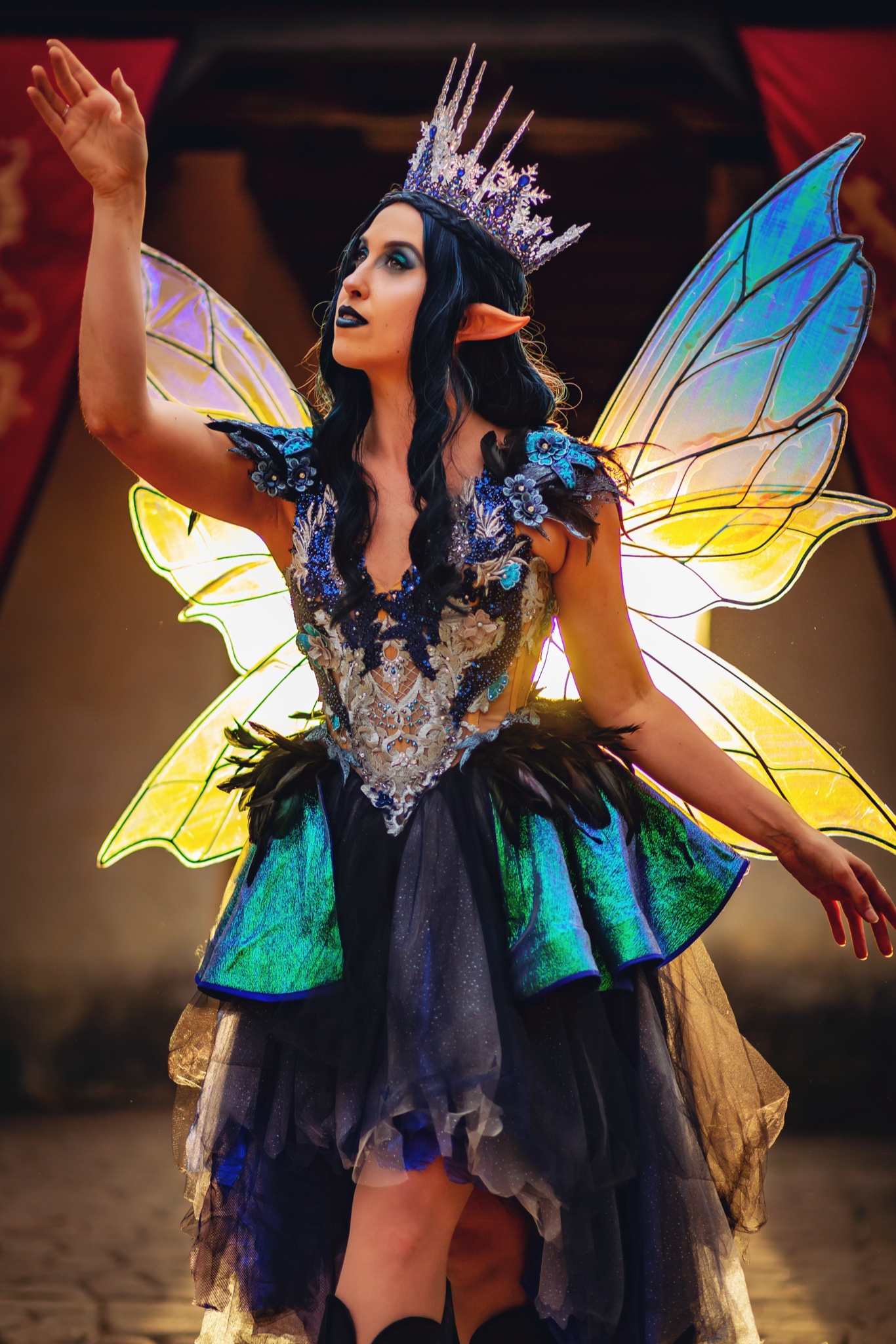 A person dressed as a fairy or queen with iridescent butterfly wings, wearing an elaborate crown, dark makeup, and a fantasy-inspired costume, performing or posing.
