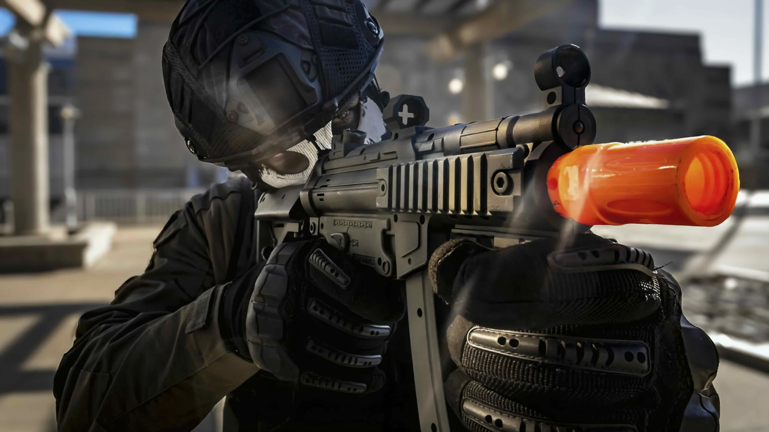 A person dressed in tactical gear aiming a firearm with an orange suppressor, wearing a helmet and gloves, outdoors in an urban area.