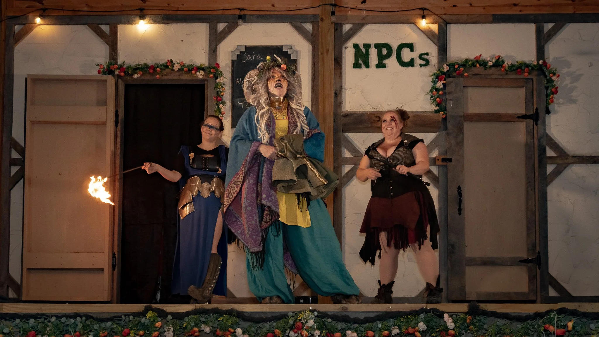 Three women in fantasy costumes performing on a decorated stage, with the central woman dressed as a wizard or witch holding a cloth, another woman in gothic armor holding a flaming torch, and a third woman in a pirate or rogue costume smiling, with 