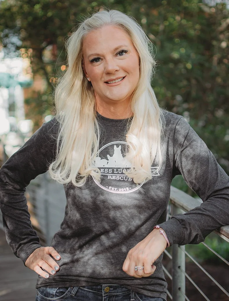 A woman with long blonde hair smiling outdoors, wearing a black and gray tie-dye sweatshirt with a rescue organization logo.