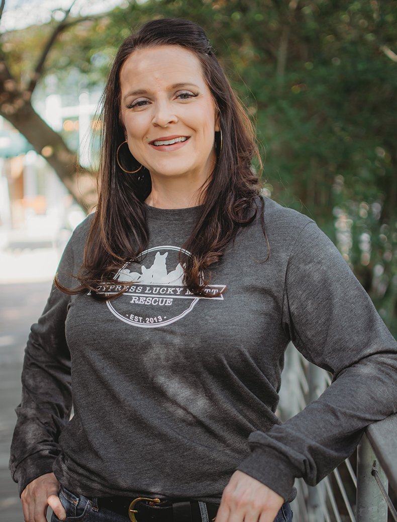 A woman standing outdoors with trees in the background. She has long dark hair, wears hoop earrings, and is smiling. She is dressed in a dark long-sleeve shirt with a rescue organization logo that includes a silhouette of a dog and the text 'Express Lucky N Rescue,' established 2013.