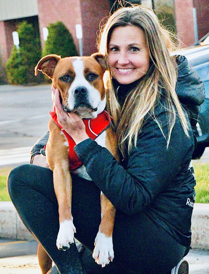 A woman with long blonde hair smiling and holding a large brown and white dog with a red collar.
