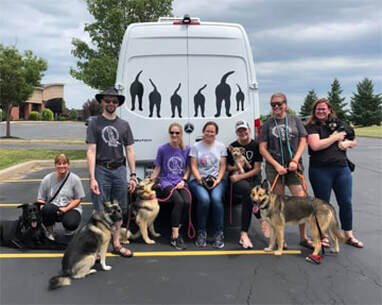Group of six people with five dogs in a parking lot, in front of a van with black dog silhouettes on it
