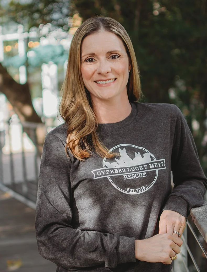A woman with long, light brown hair smiling, wearing a gray sweatshirt with 'Cypress Lucky Mutt Rescue' logo, outdoors with trees in the background.