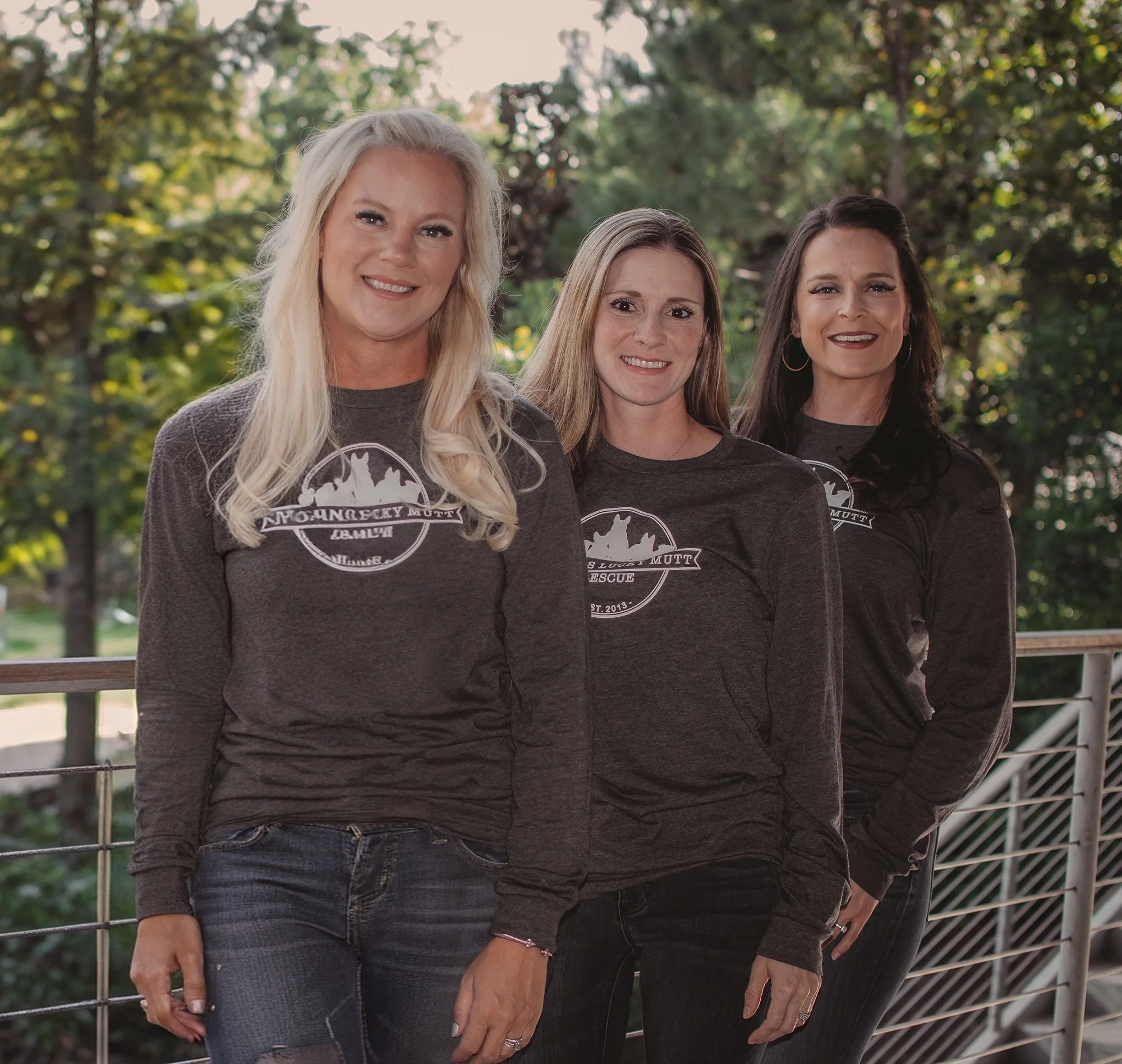 Three women standing outdoors, smiling, wearing matching dark gray long-sleeve shirts with a logo and text, posing on a balcony with trees in the background.