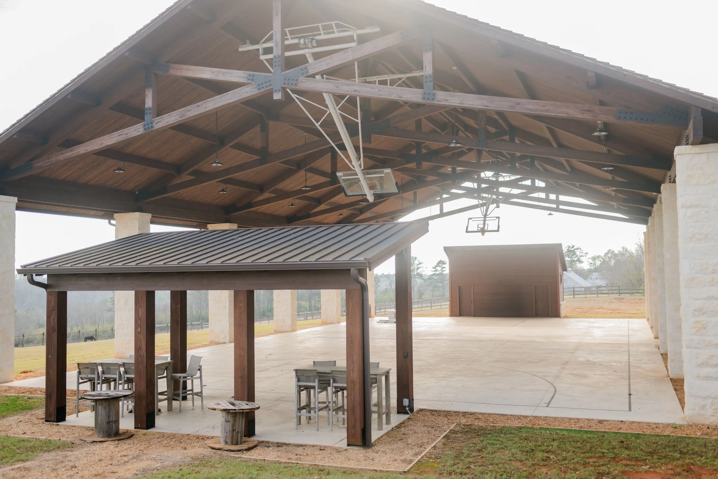An outdoor covered pavilion with a basketball court underneath and a smaller shelter with outdoor tables and chairs.
