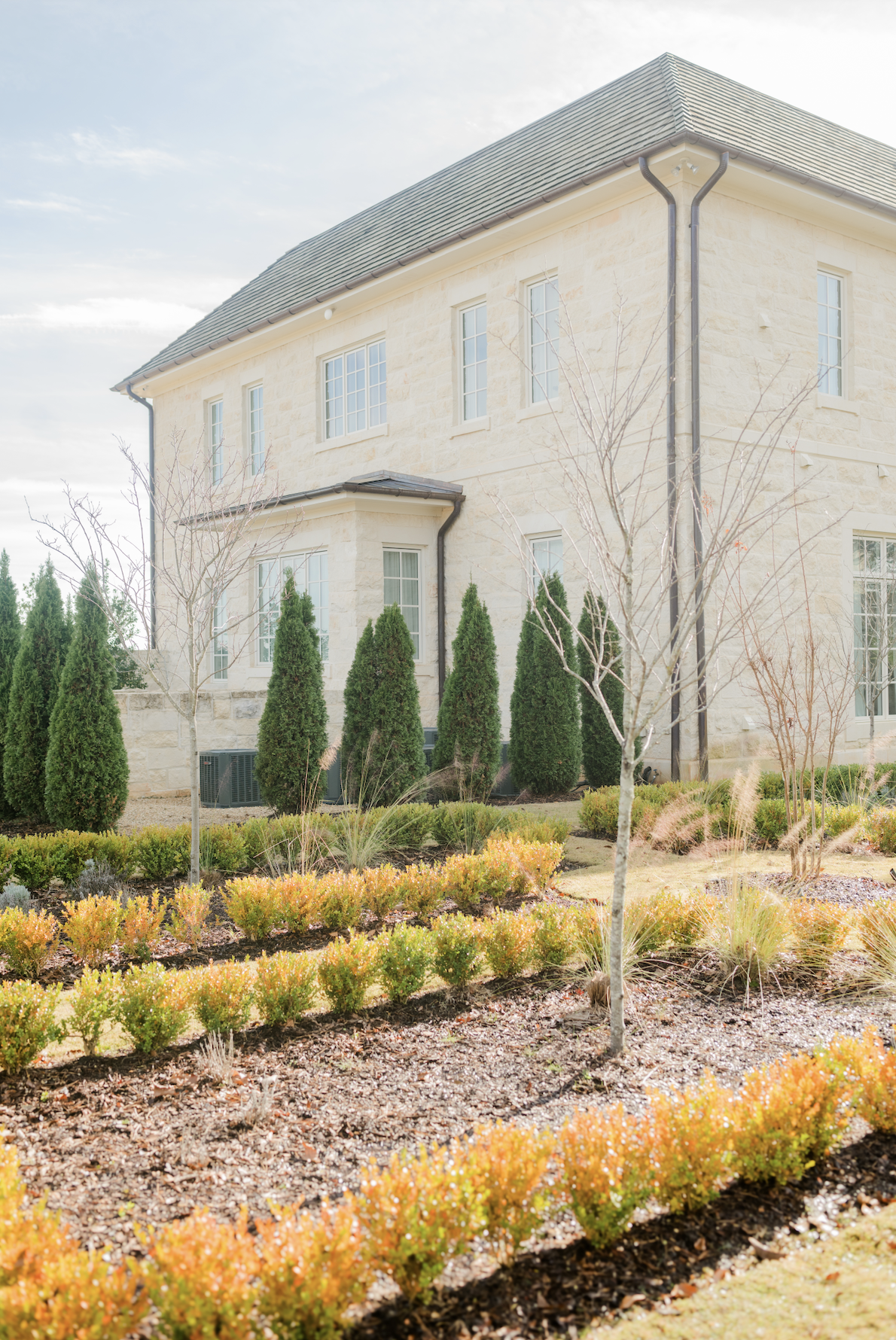 Front view of a beige stone house with large windows and a dark gray roof, surrounded by landscaped bushes and small trees.