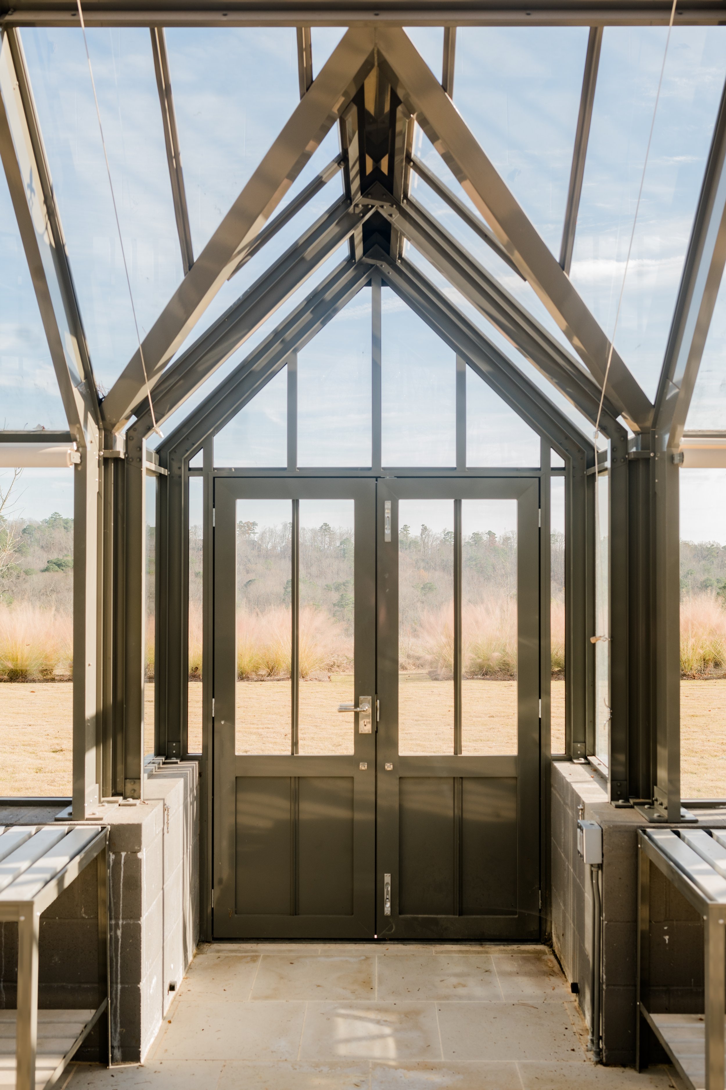 Interior view of a modern greenhouse with glass walls and ceiling, showing an outdoor landscape of grass and trees through the glass.