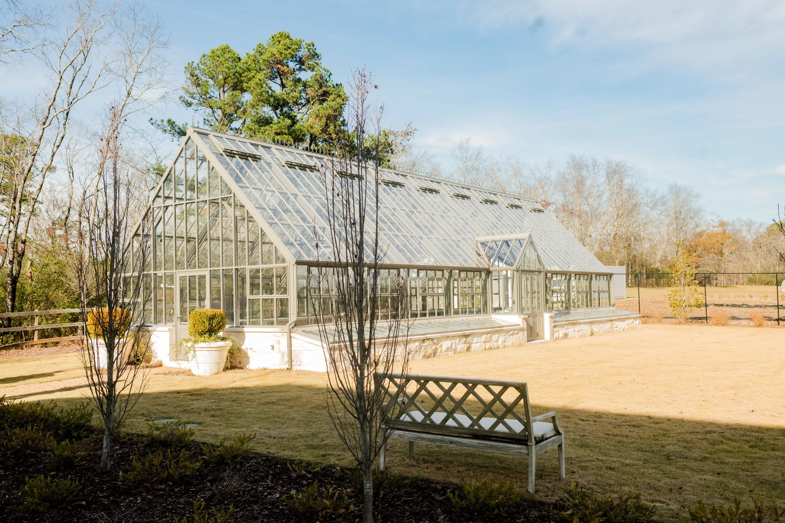 A large glass greenhouse with a peaked roof and multiple windows, surrounded by a yard with grass, trees, and a wooden bench.