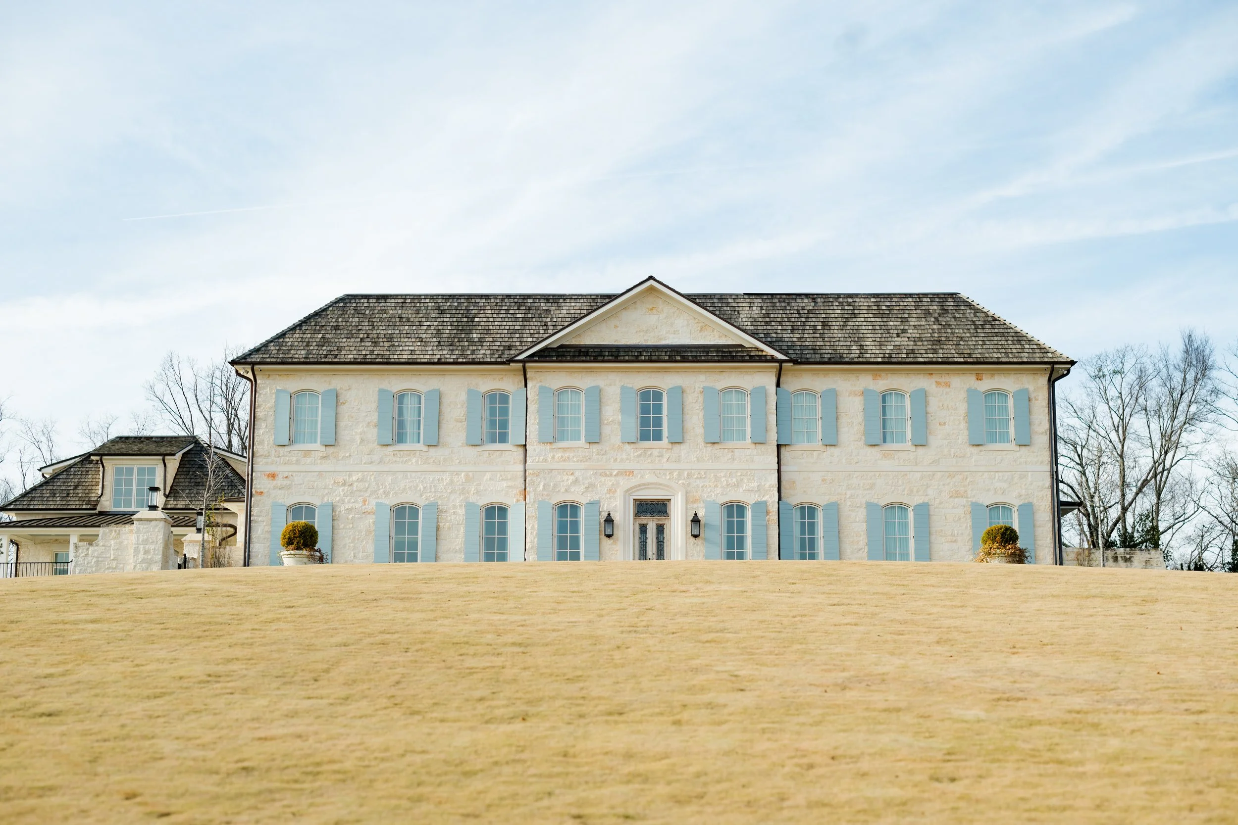 A large, two-story beige house with blue shutters and a dark shingled roof, situated on a barren grassy hill under a partly cloudy sky, with leafless trees in the background.