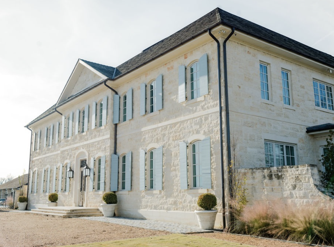 A large, elegant house with white stone walls, blue shutters, and multiple windows, featuring a gravel driveway and potted plants outside.