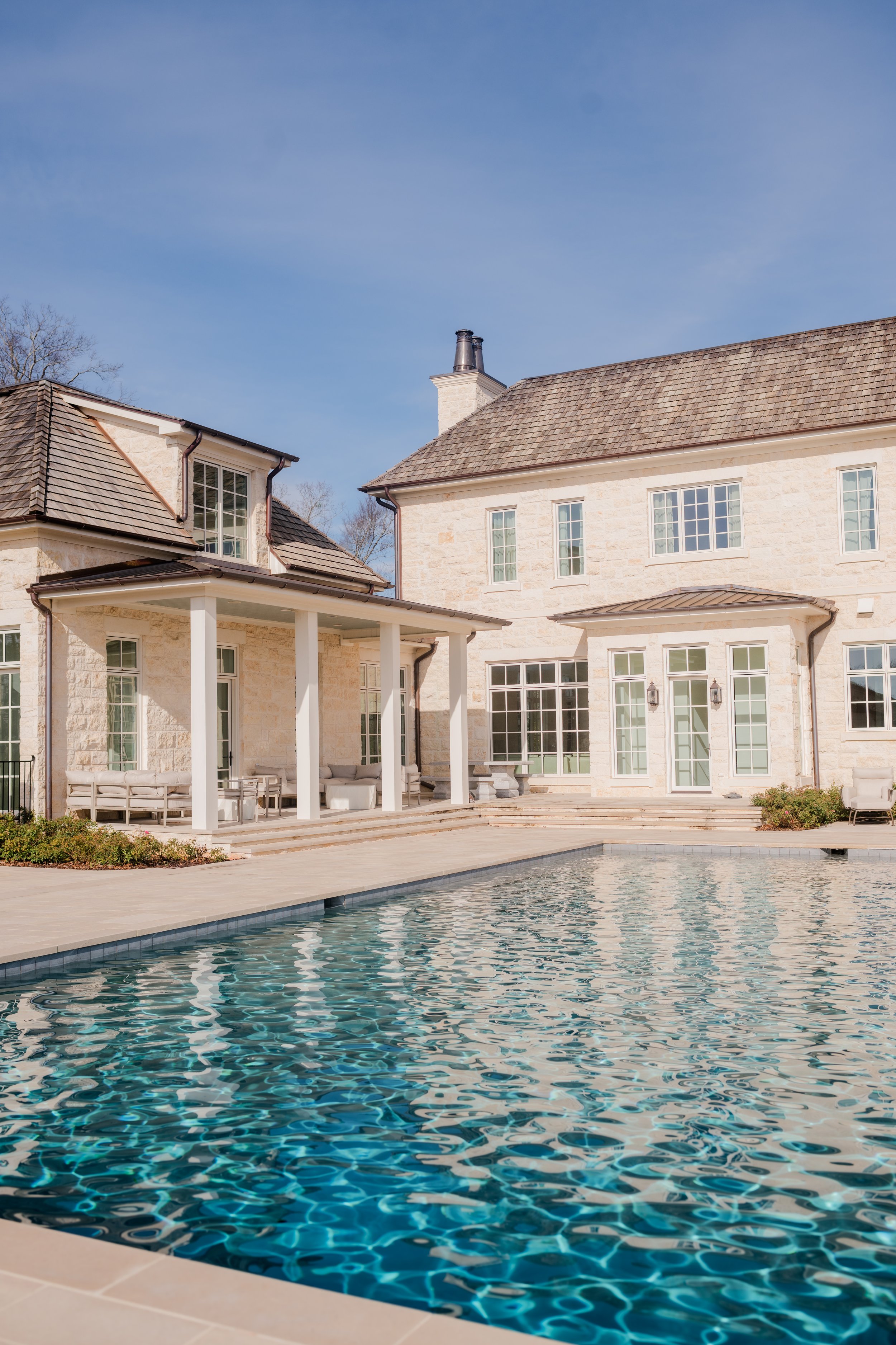 Luxury house with a swimming pool, stone exterior, large windows, covered patio area with seating, and a clear blue sky.