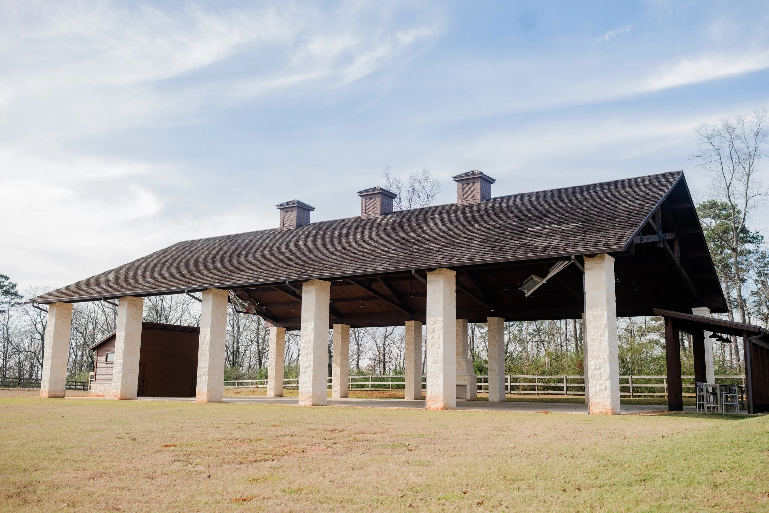 A large outdoor pavilion with stone columns, a shingled roof, and open sides, situated in a grassy area with trees in the background under a partly cloudy sky.