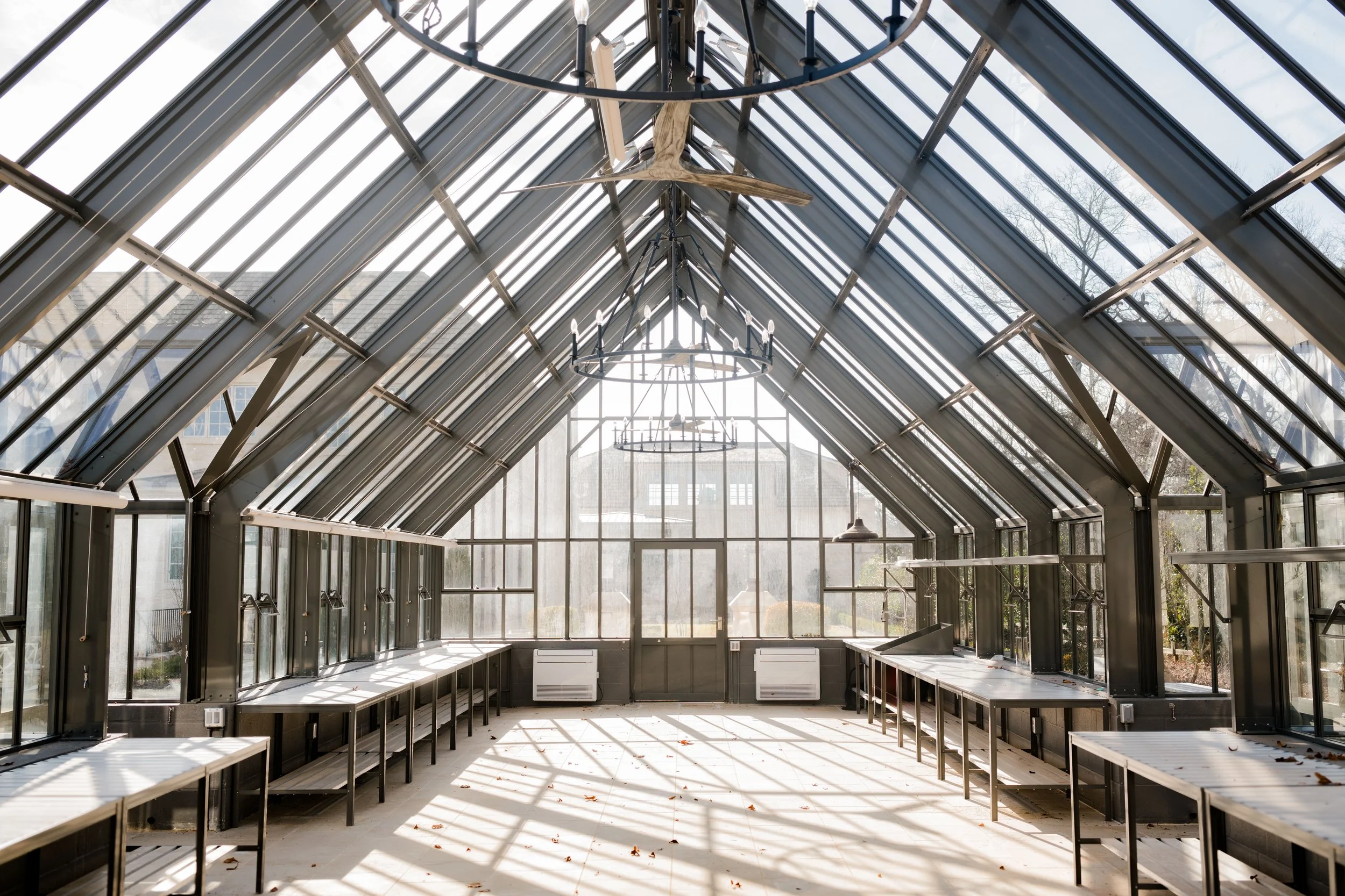 Sunlit greenhouse interior with glass walls and ceiling, metal framework, wooden benches along sides, chandelier hanging from ceiling, and a ceiling fan.
