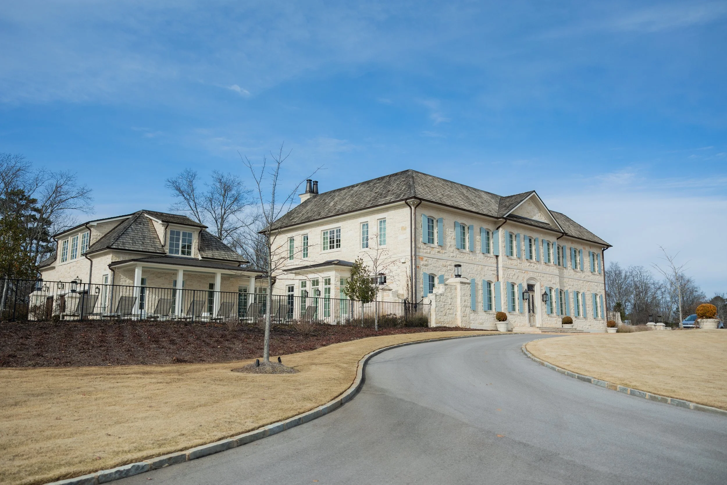 Large, elegant mansion with light-colored stone exterior, multiple windows with blue shutters, and a curved driveway leading to its entrance, surrounded by a landscaped yard and some leafless trees under a blue sky.