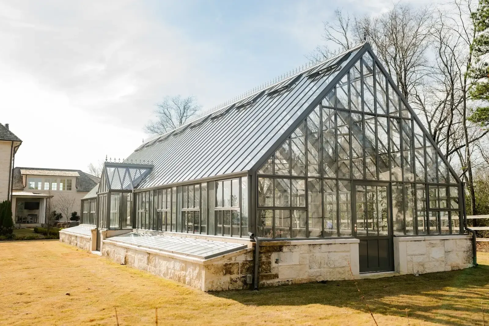 A large glass greenhouse with a metal frame, situated on a stone foundation, in a backyard with surrounding trees and neighboring houses.