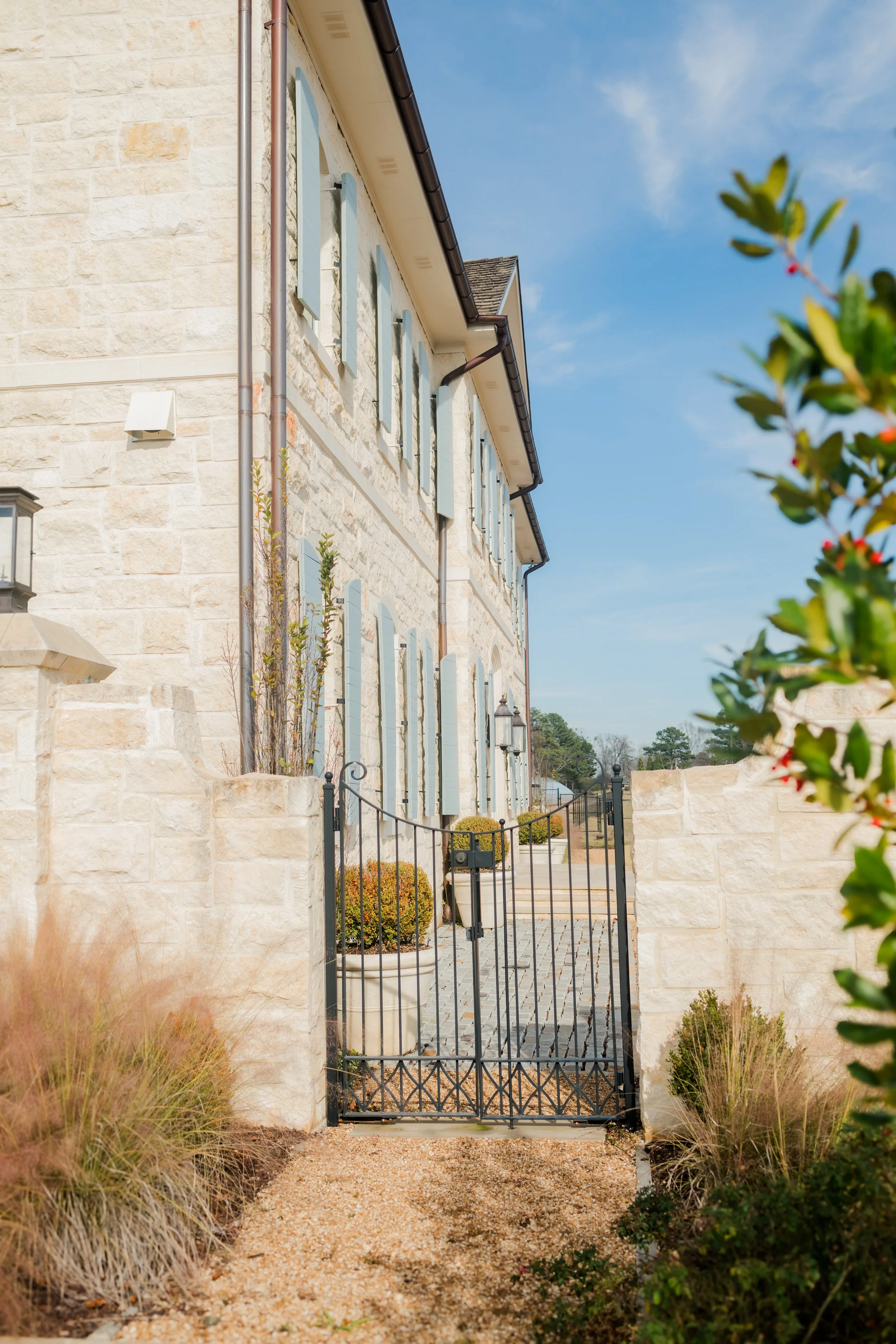 A photo of a gated entrance leading to a stone house. The house has multiple blue shutters on its windows and decorative plants in pots. The sky is clear and blue.
