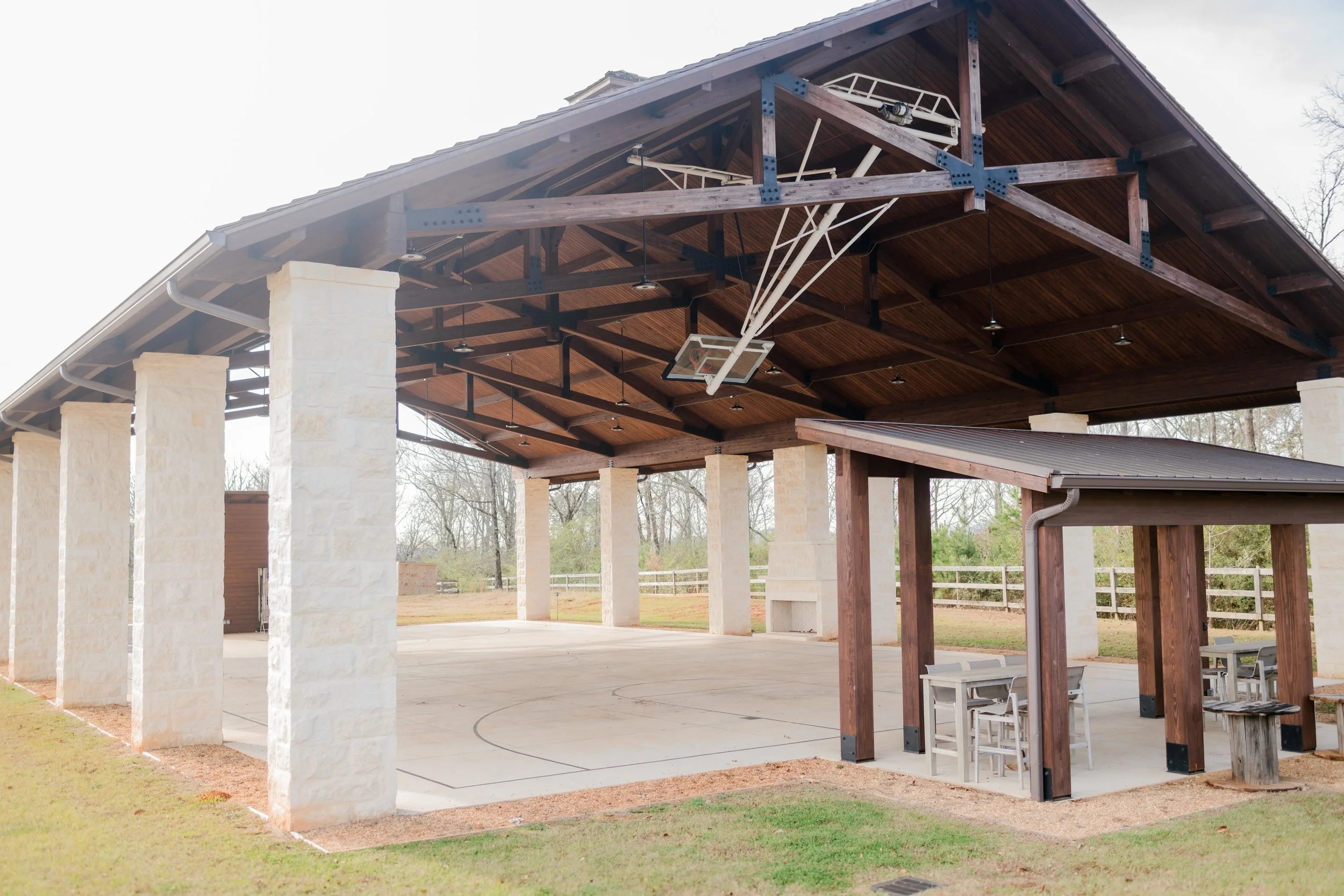 Covered outdoor basketball court with a wooden roof, stone pillars, and a small shaded seating area with tables and chairs.