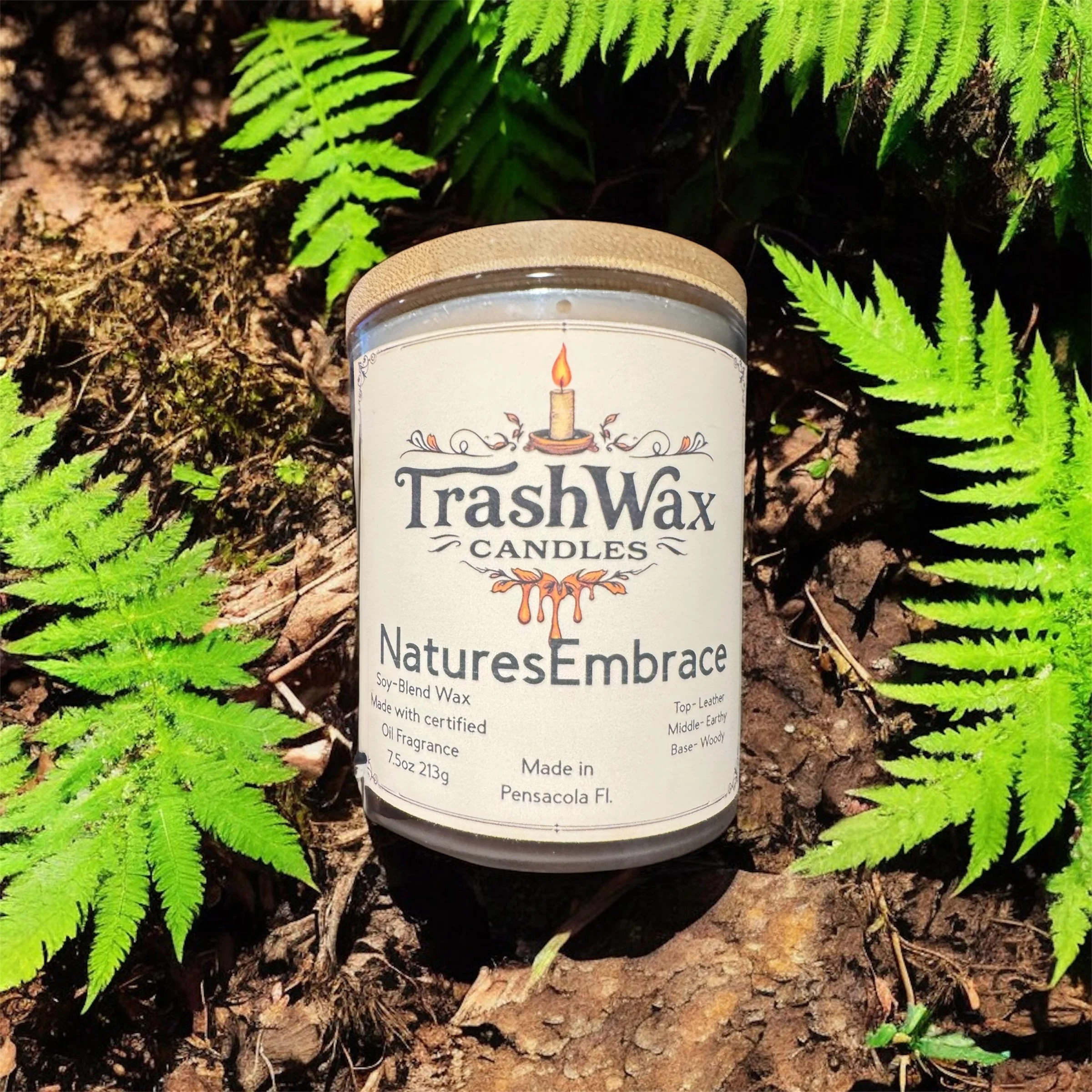 A candle named 'TrashWax Candles NaturesEmbrace' resting on the ground among green ferns and soil. The label indicates it is made from soy-blend wax with a woodsy scent, and it is made in Pensacola, Florida.