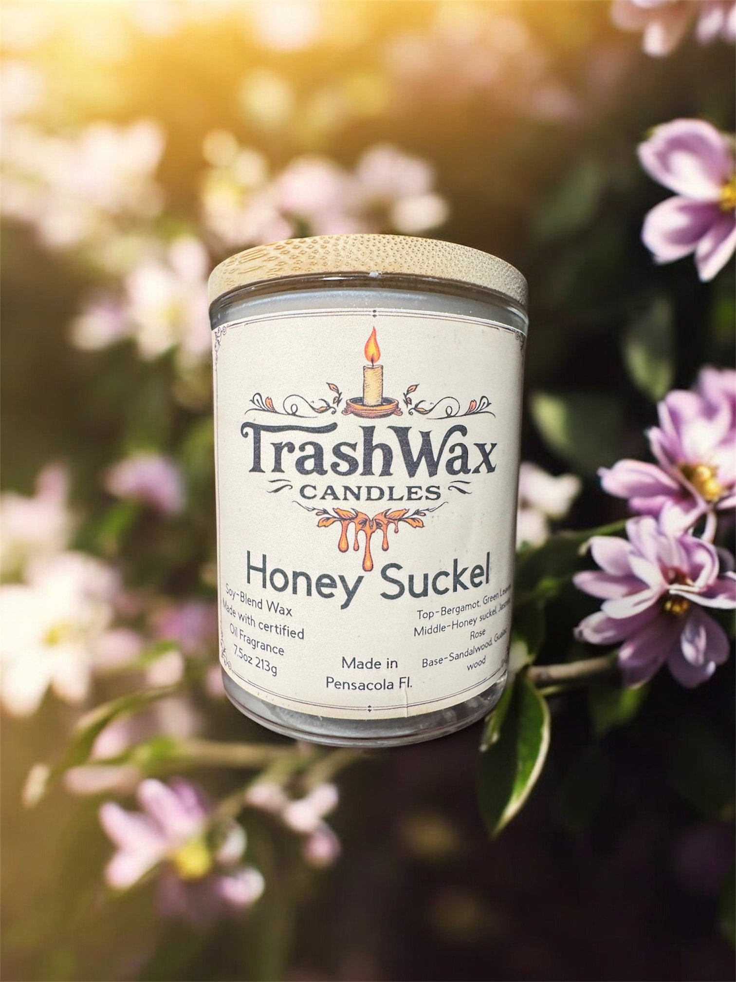 A jar of Trash Wax Candles Honey Suckle scent surrounded by pink and purple flowers, with a blurred natural background and sunlight.