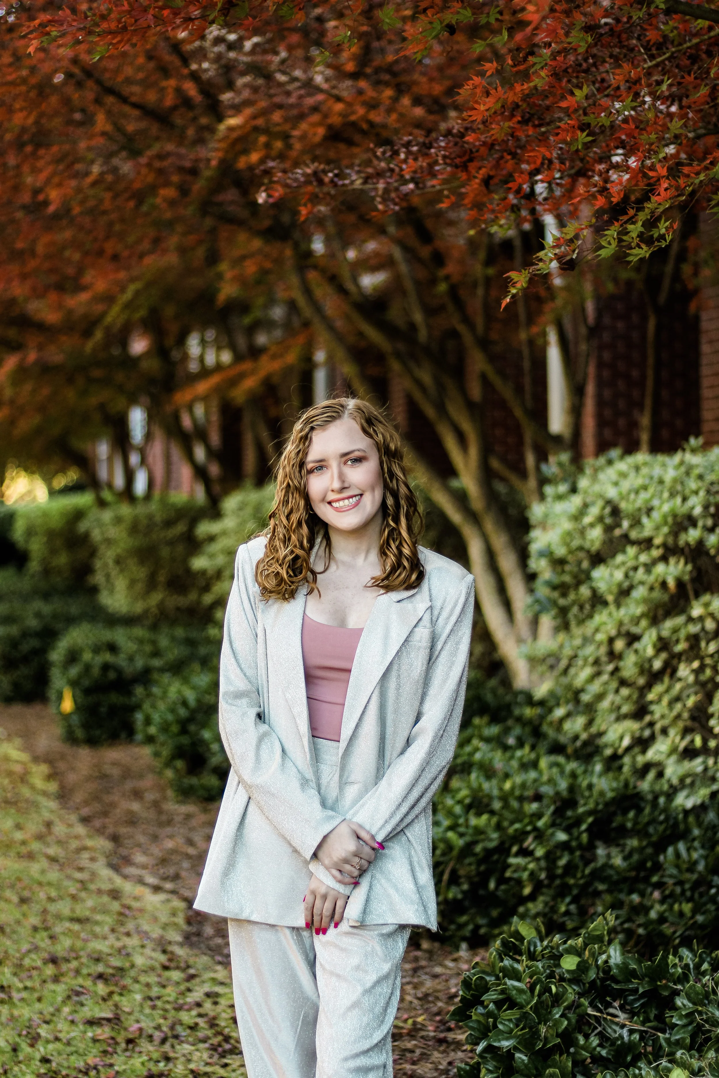 A young woman with curly red hair and a big smile posing outdoors, wearing a light-colored blazer and matching pants, with a pink top underneath. She is standing on a garden path with dense bushes and trees with red leaves in the background.