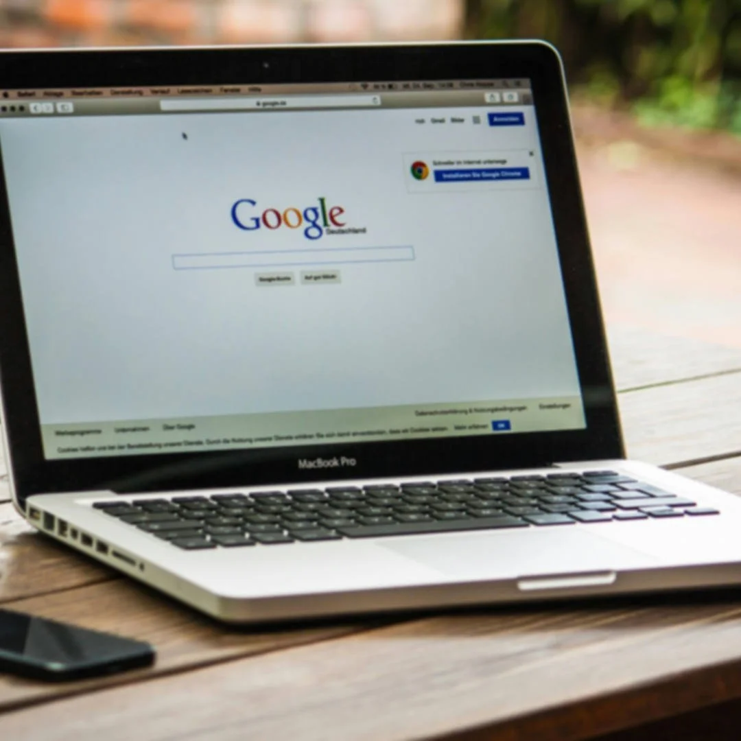 Open MacBook Pro displaying Google search page on a wooden table outdoors with a smartphone nearby.