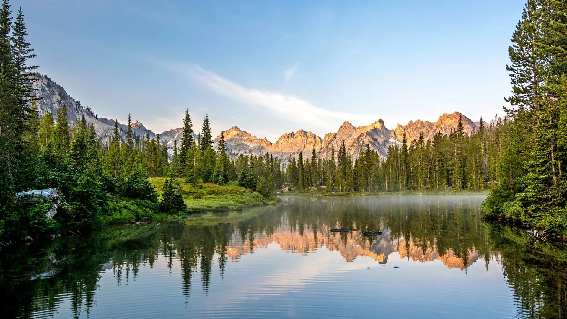 A serene lake surrounded by dense evergreen trees with mountain peaks in the background, during sunrise or sunset with a light mist over the water.
