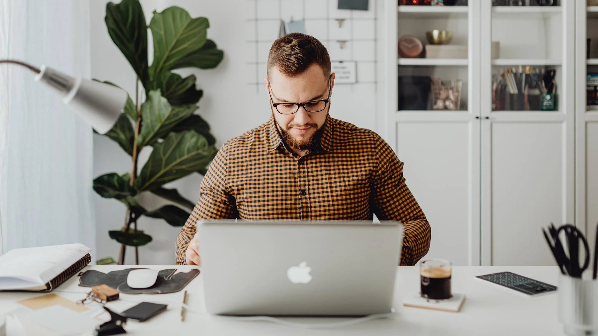 A man with glasses and a beard working at a desk with a laptop, coffee, and office supplies in a modern, organized office with white cabinets and a large green plant.
