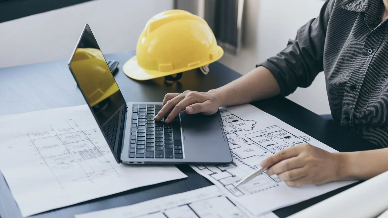 Person analyzing architectural blueprints with a laptop and a yellow safety helmet on the desk.