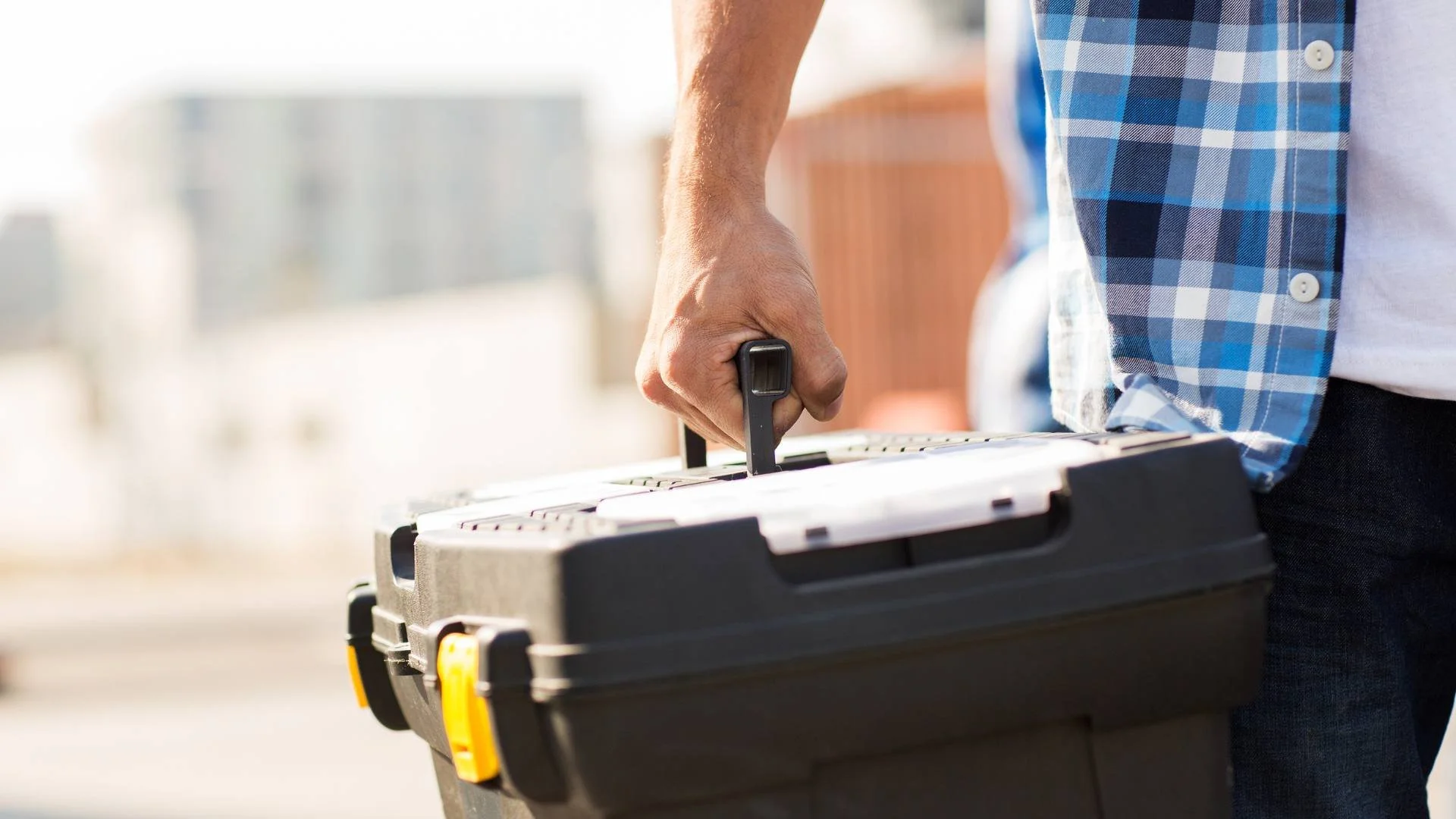 Person wearing a blue checkered shirt holding a black toolbox outdoors