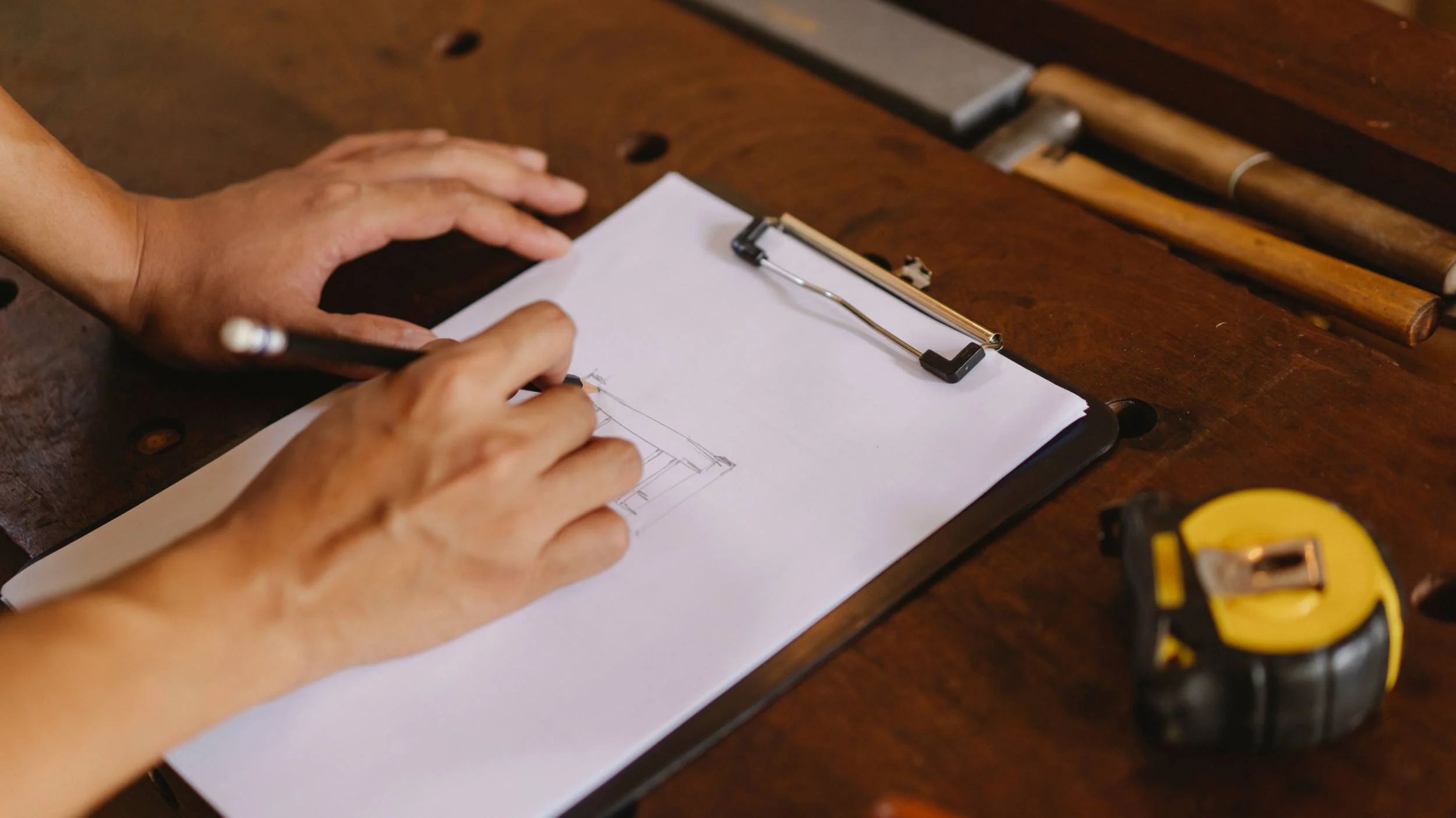 Person sketching a building on a clipboard with a pencil, surrounded by measuring tape and construction tools on a wooden workbench.