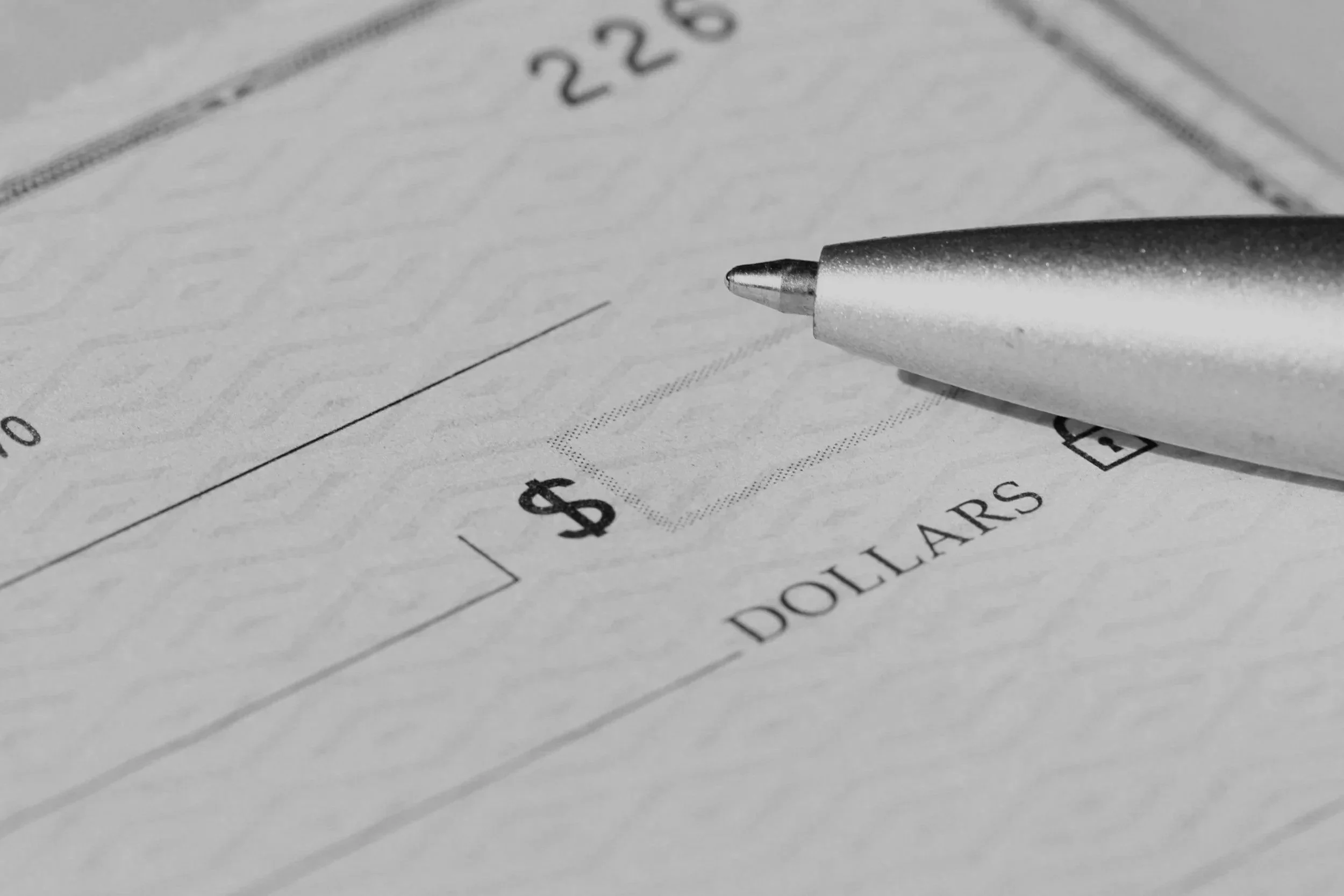 Close-up of a silver pen resting on a check or bank document with dollar signs, numbers, and the word 'DOLLARS' in black and white.