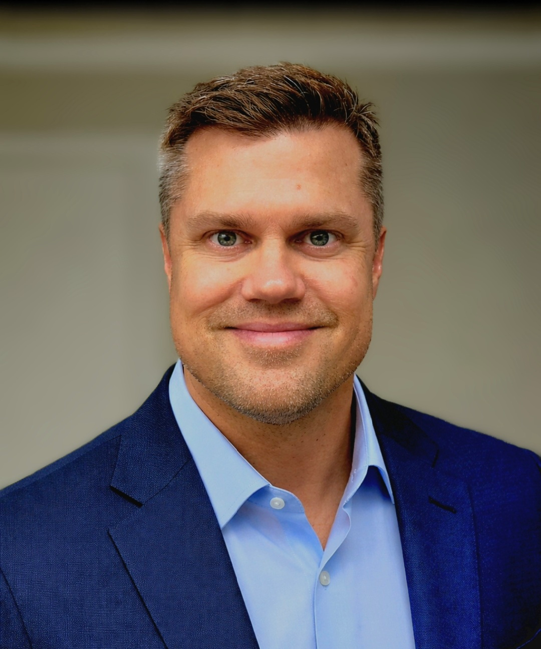 Headshot of a smiling man with short brown hair, wearing a blue suit jacket and light blue collared shirt, against a neutral background.
