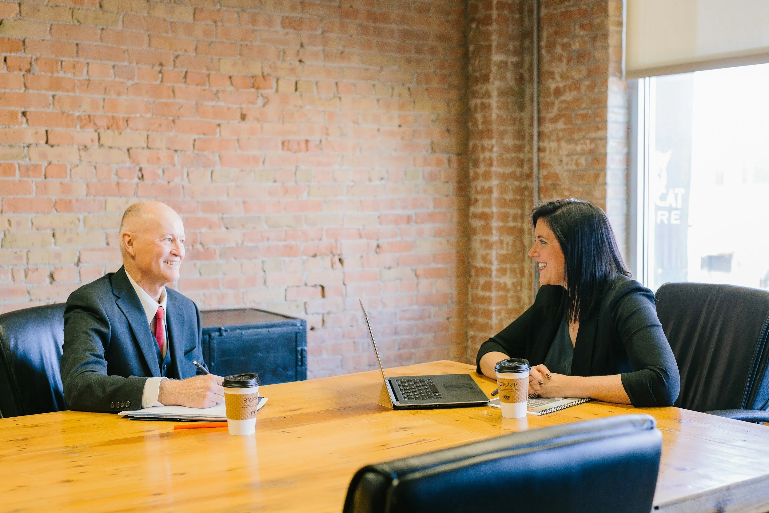 A man and a woman are sitting at a wooden table in an office with exposed brick walls, having a conversation. Both are smiling. The man has short blond hair and is wearing a suit, while the woman has long dark hair and is wearing a black blazer. There are two coffee cups, a laptop, and some notebooks on the table. Large windows are in the background.