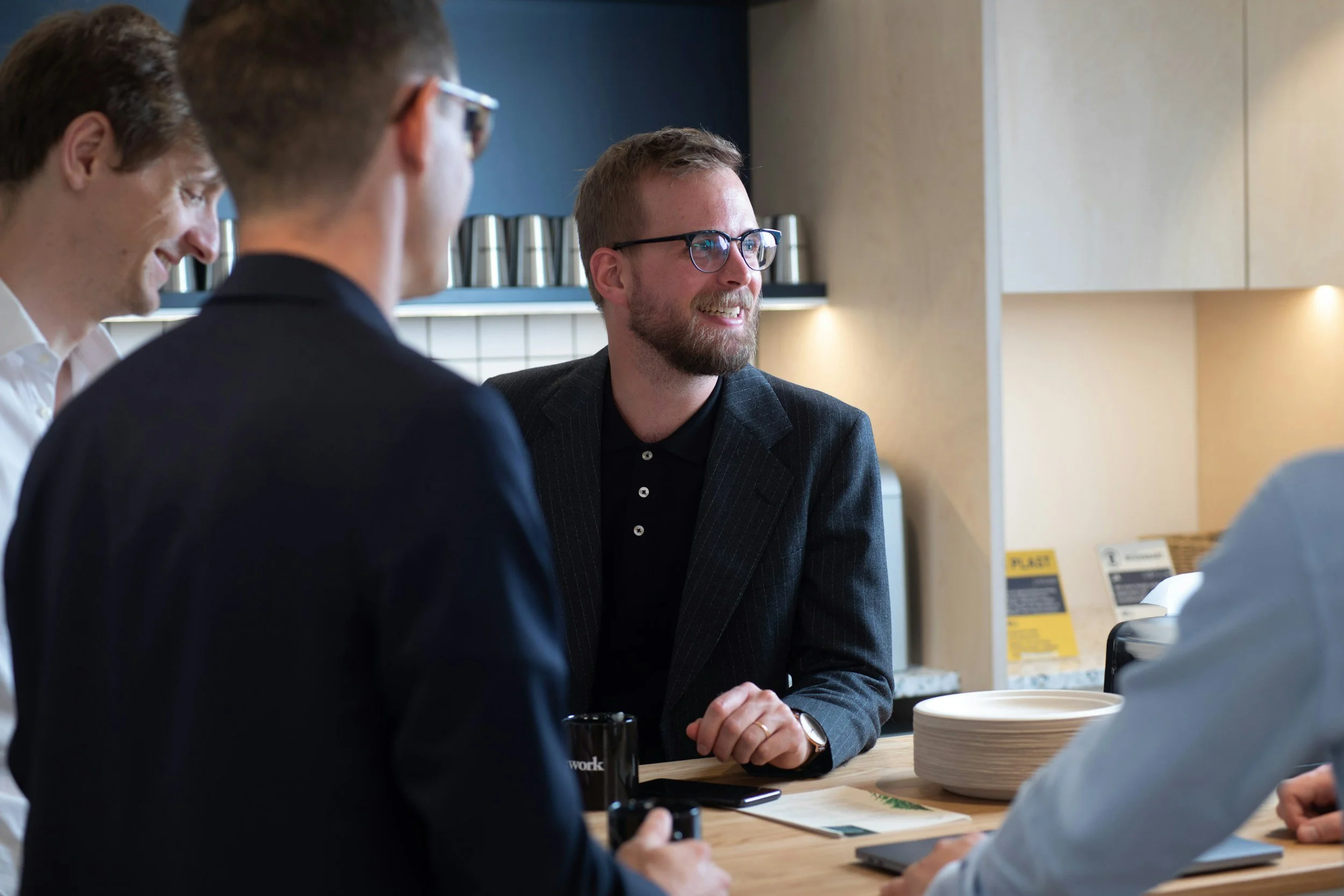 A group of four men having a discussion in a modern office space with light wood accents and shelves. The man in glasses and a dark blazer is smiling and engaged in conversation.