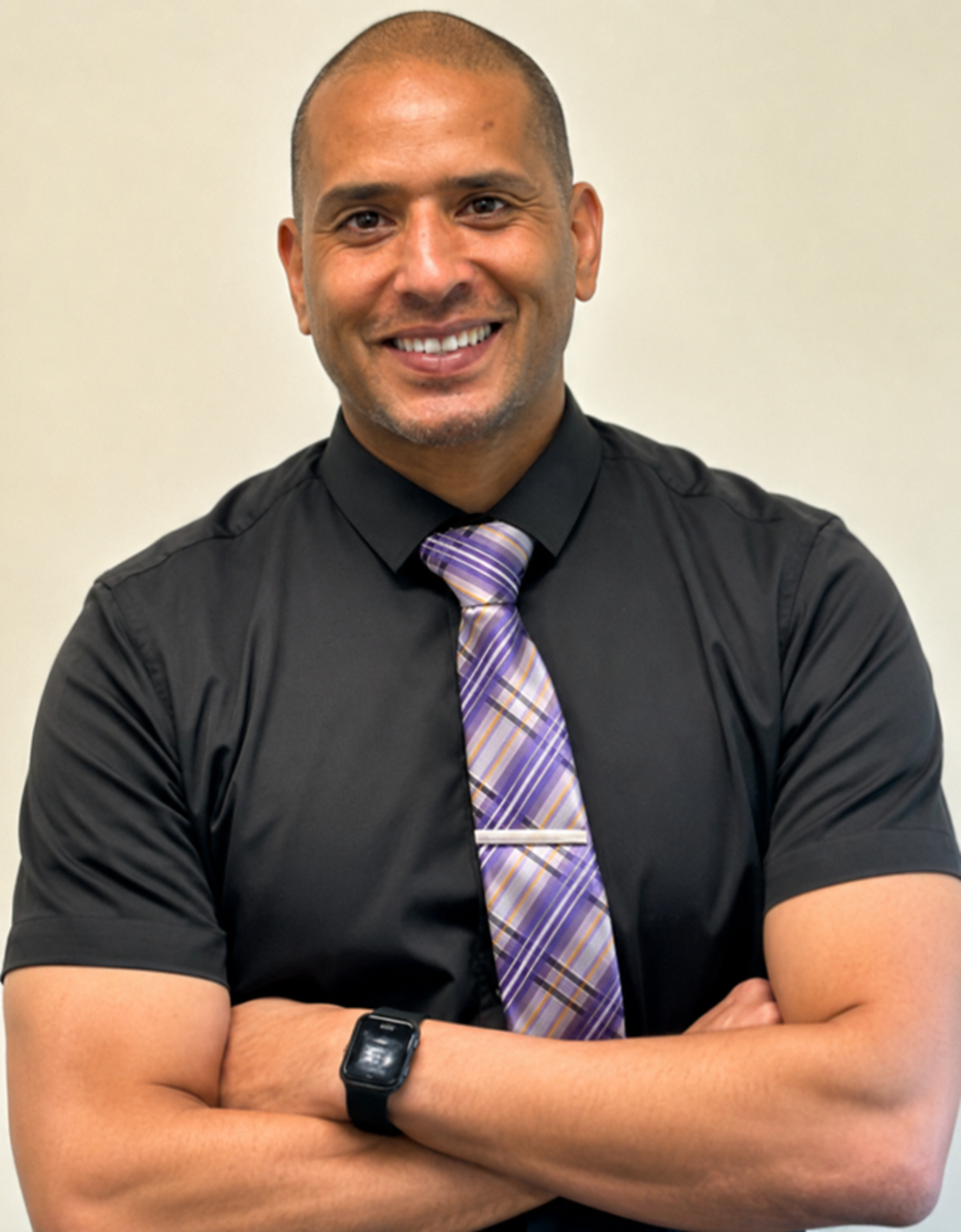 A smiling man with a shaved head wearing a black shirt, a purple patterned tie, and a black watch, standing with arms crossed against a plain beige background.