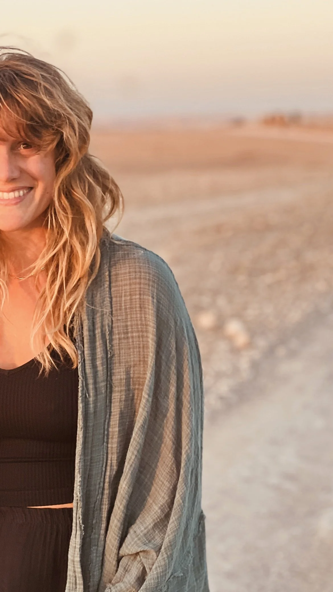 A smiling woman with wavy blonde hair standing on a beach at sunset, wearing a black top and a semi-sheer, plaid button-up shirt.