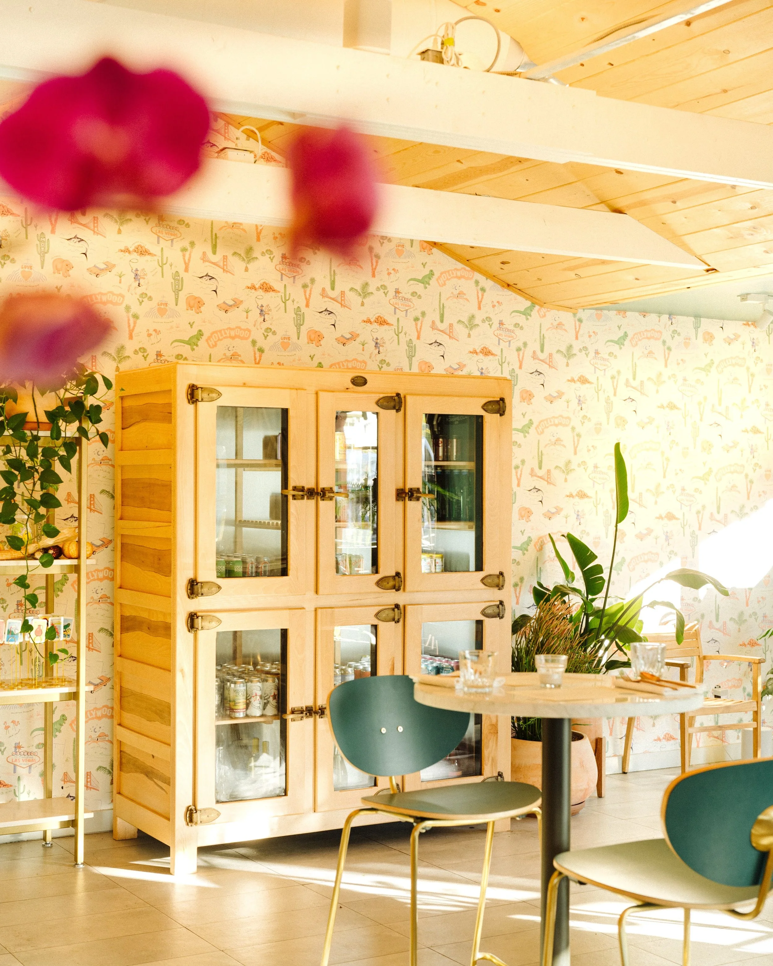 Cozy interior of a cafe or restaurant with a wooden cabinet, a table with glasses, plants, and colorful wallpaper with a desert and Hollywood theme.