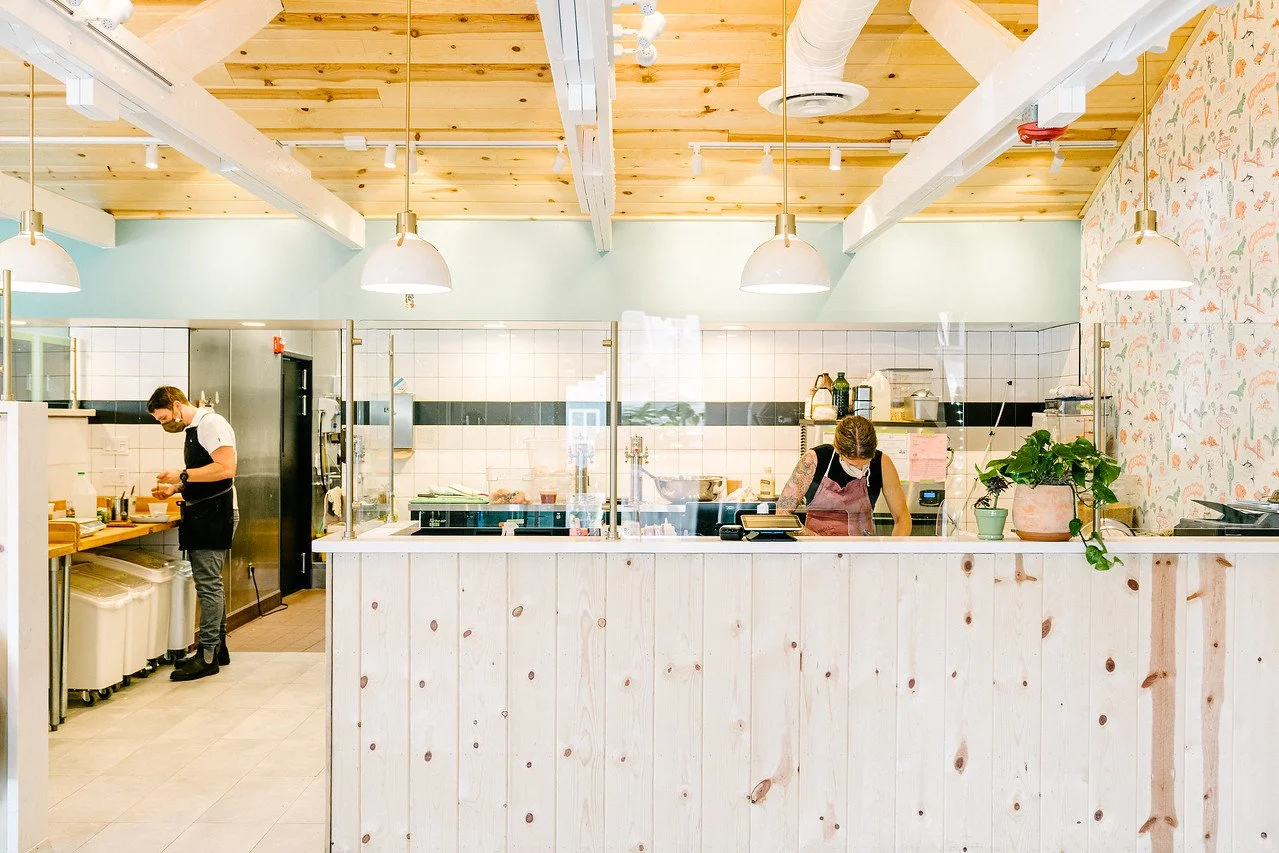 Two people working behind a wooden counter in a bright, modern kitchen or cafe, with white tiles, light green walls, hanging pendant lights, and a potted plant on the counter.