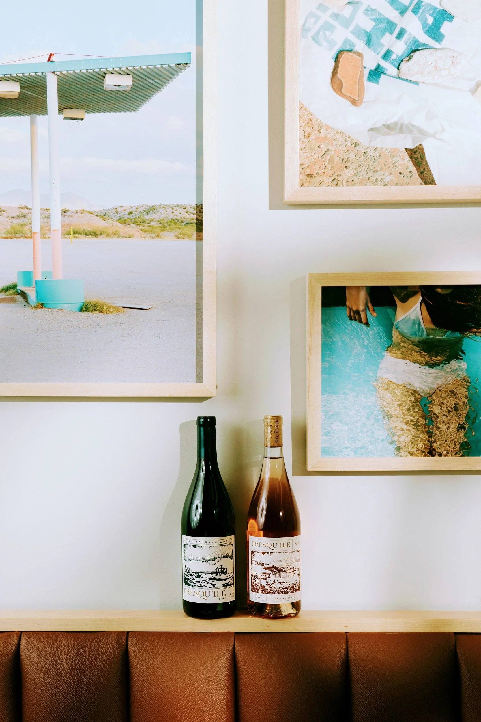 Two bottles of wine on a wooden ledge with framed artwork and photographs hanging on the white wall behind it.