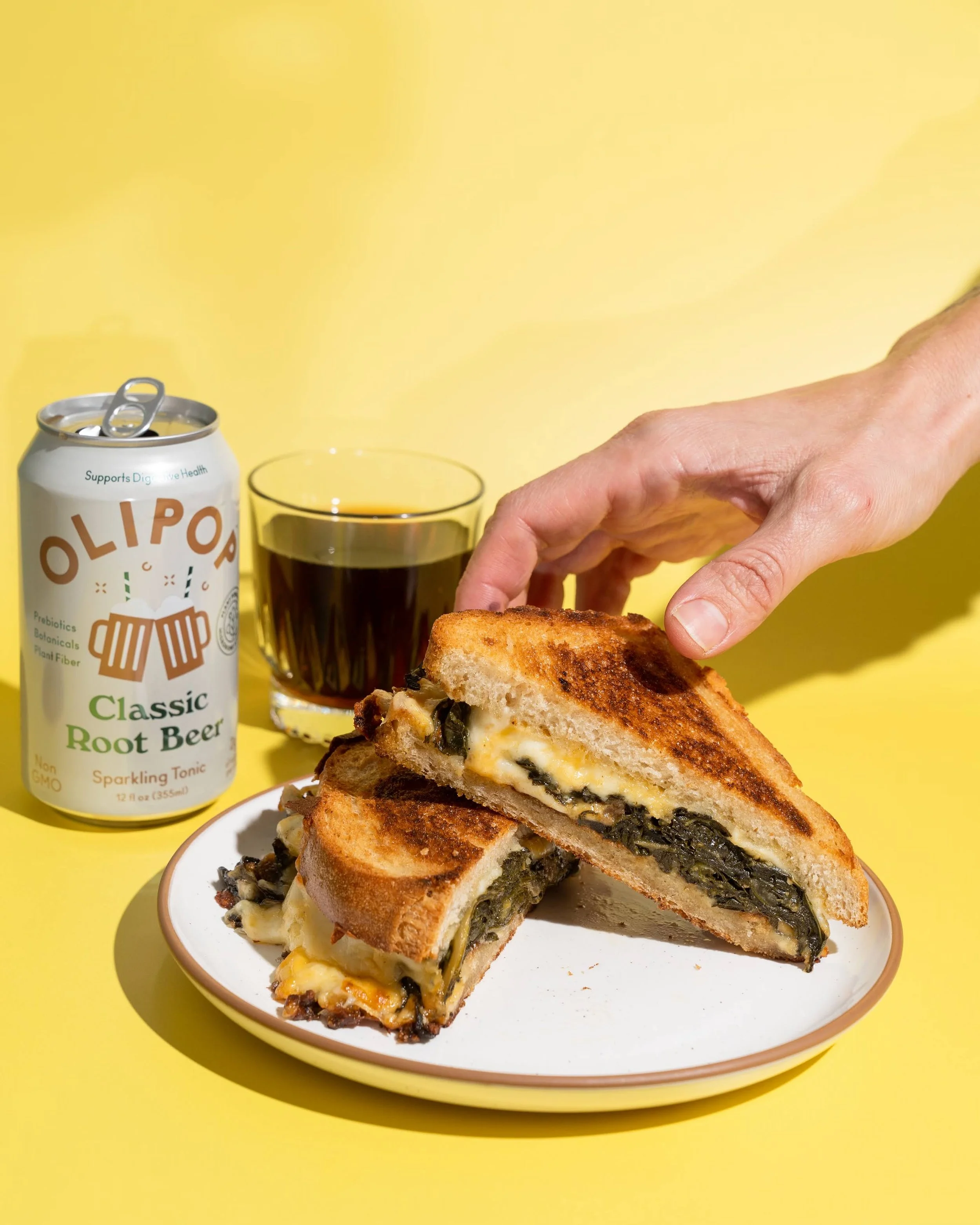 A grilled sandwich filled with leafy greens and melted cheese, a can of OLIPOP soda, and a glass of dark soda on a yellow background.