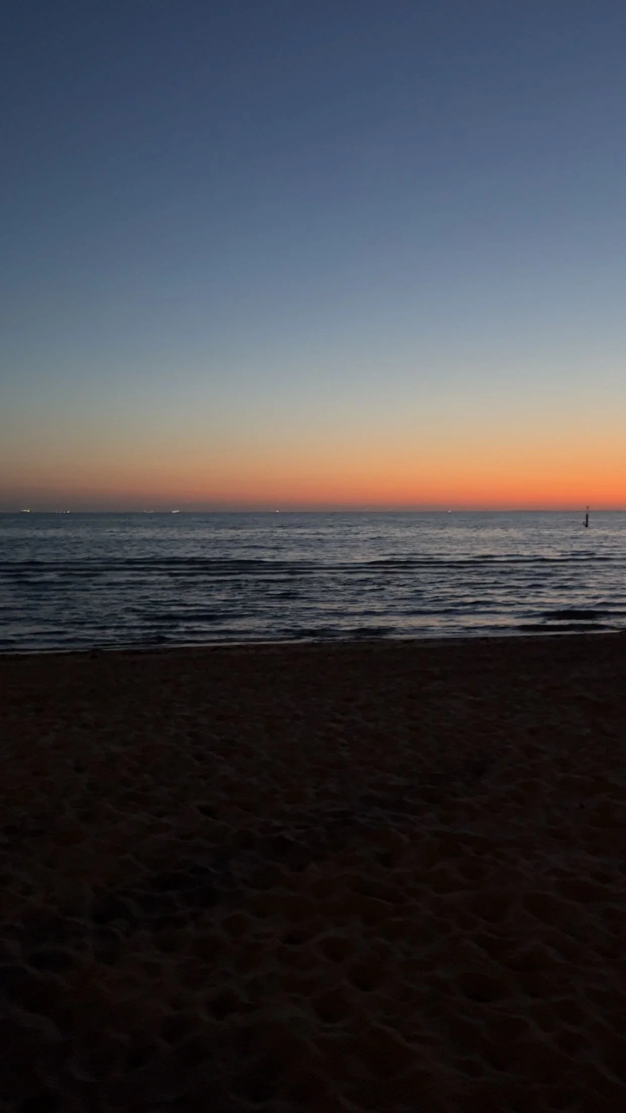 A sunset over the ocean with a clear sky and gentle waves, viewed from a sandy beach.
