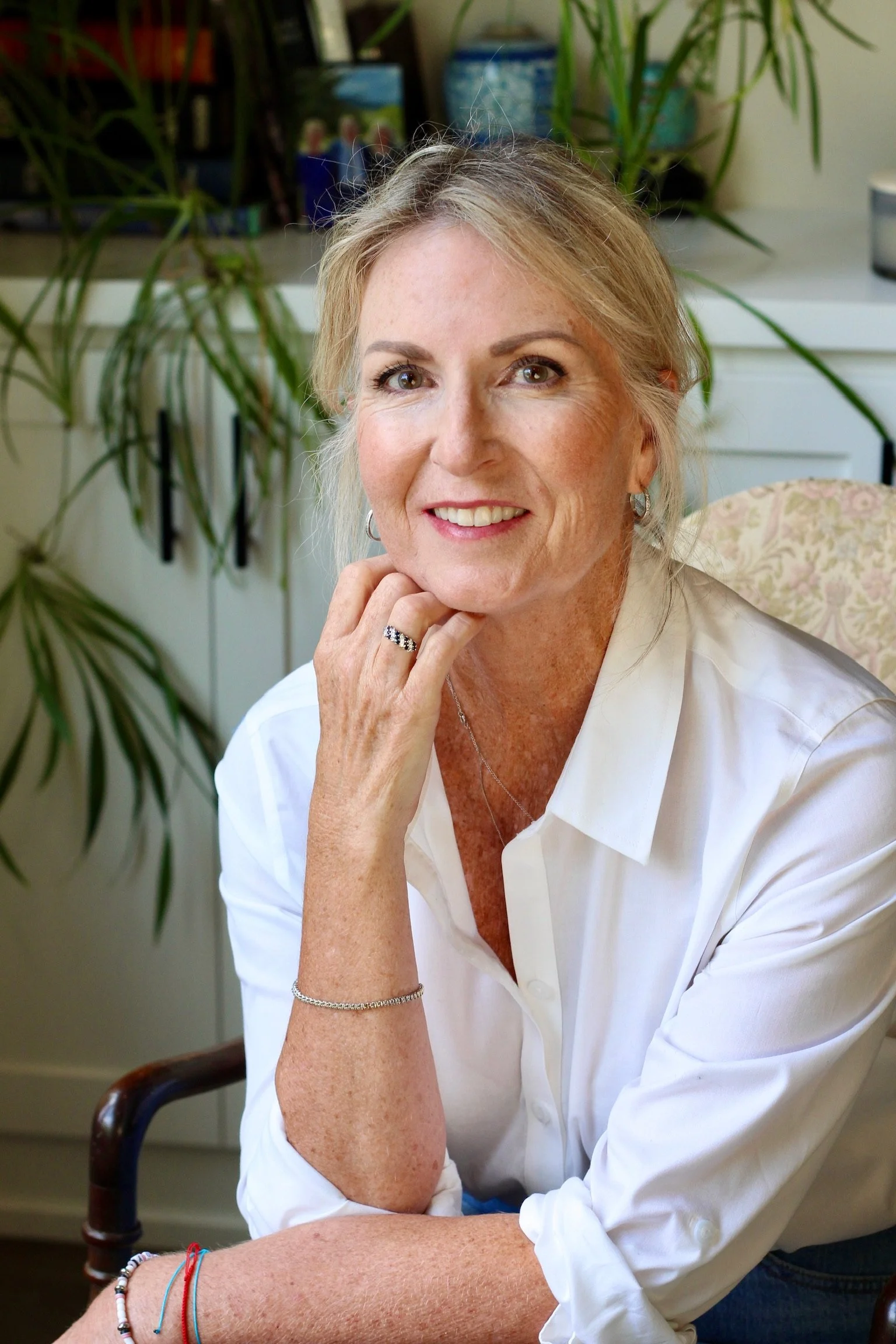 A woman with blonde hair, wearing a white blouse, sitting at a desk with a thoughtful smile, touching her chin with her right hand, in an office or home workspace with plants and shelves in the background.