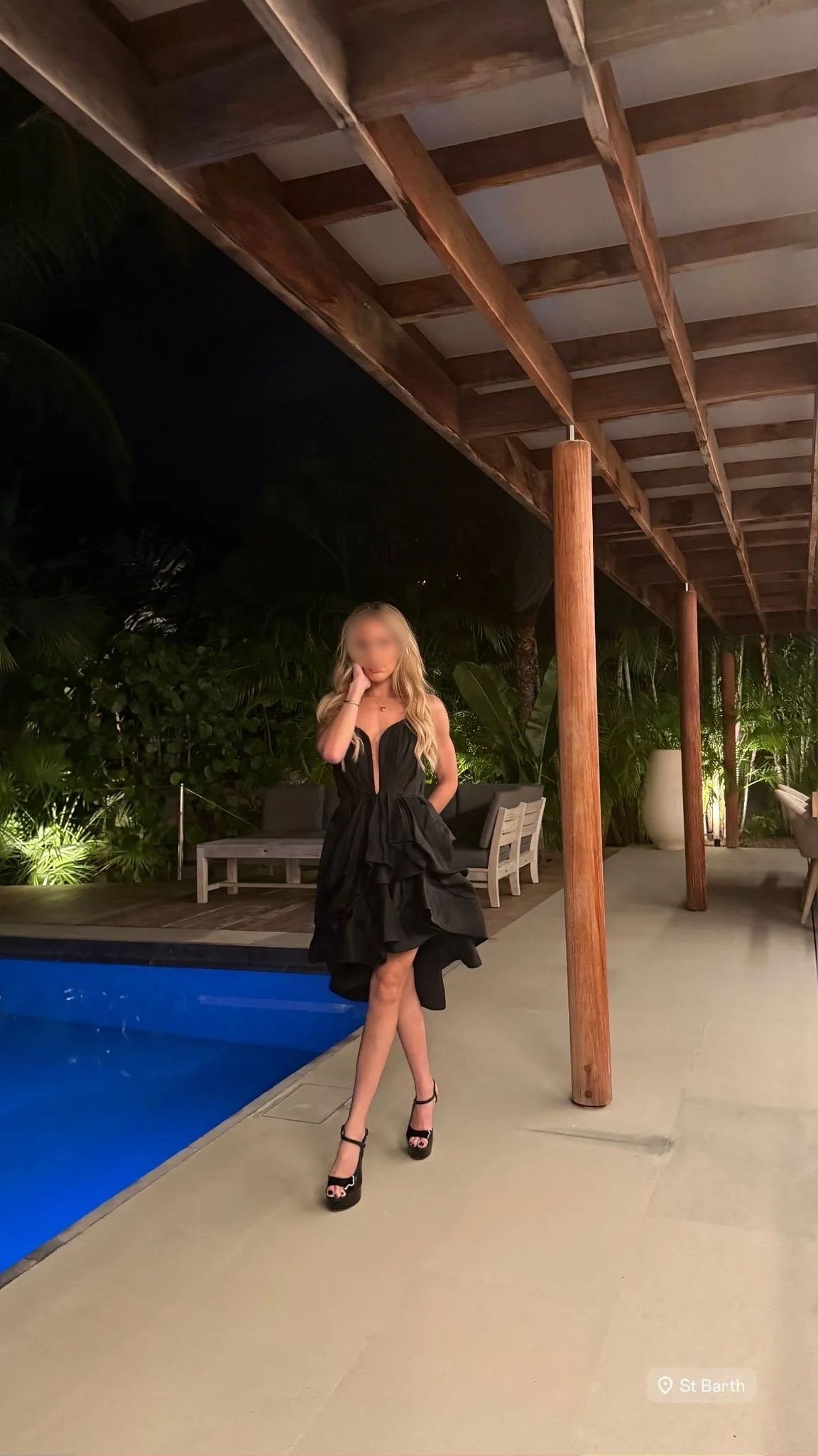 A woman with long blonde hair wearing a black dress and high heels standing beside a swimming pool at night, with lush greenery and outdoor furniture in the background.