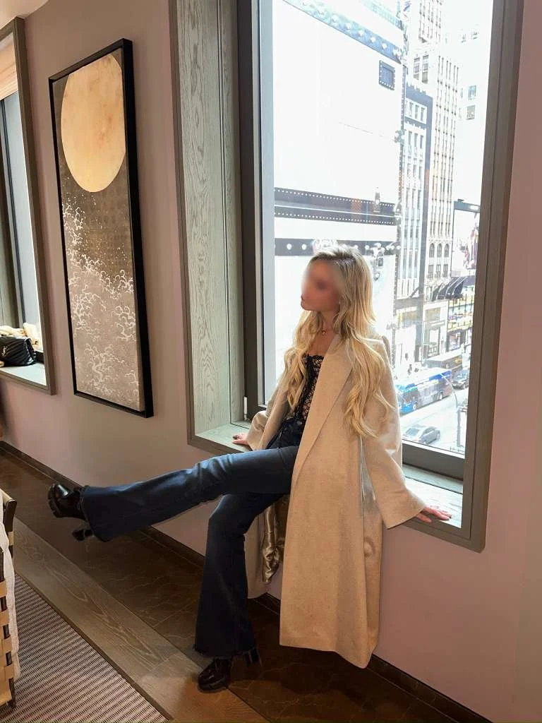A woman with long blond hair, dressed in a beige coat, black boots, and dark pants, sitting on a window sill in an urban setting, looking outside at city buildings and streets.