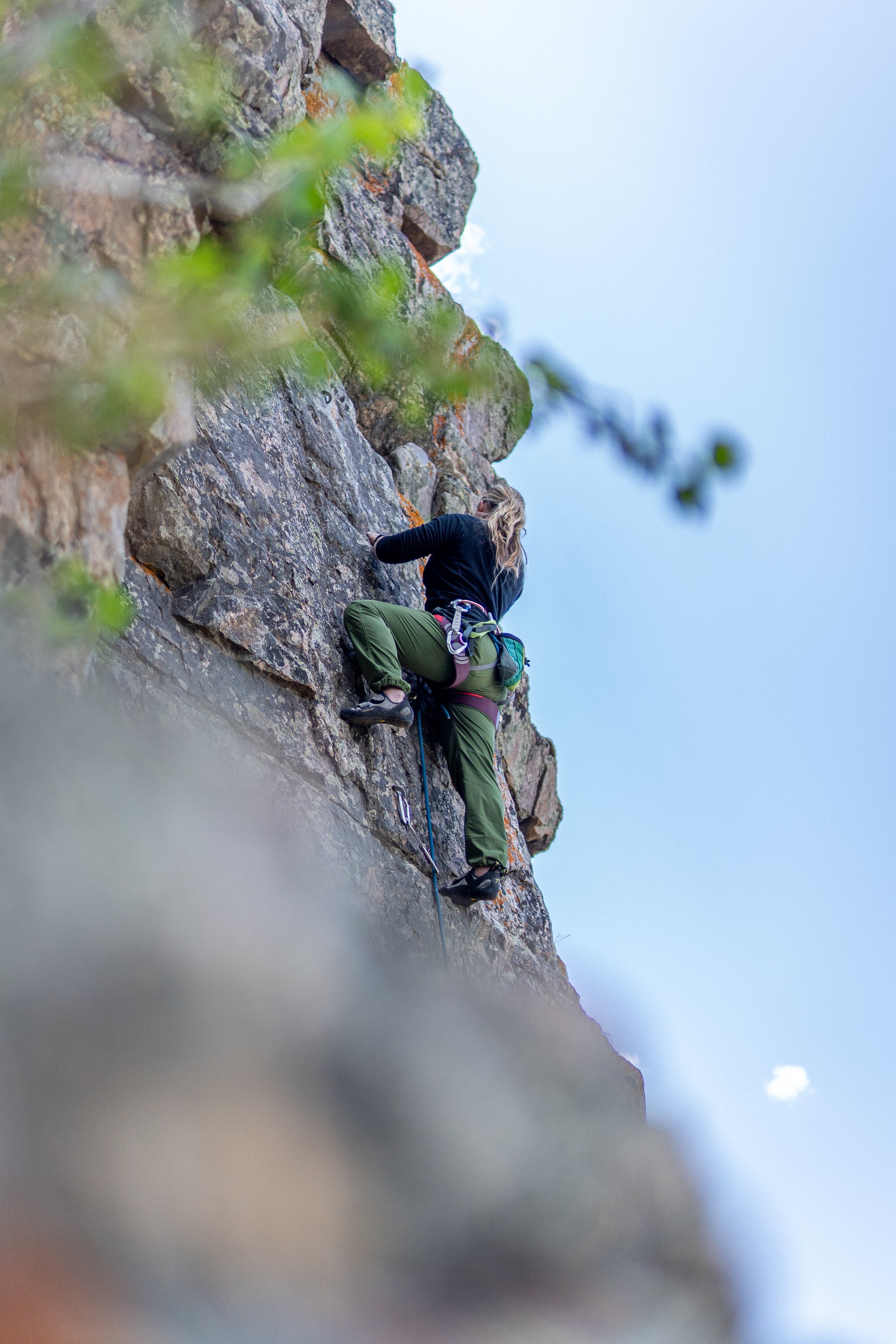 A woman rock climbing on a steep outdoor cliff with some green foliage in the foreground and a partly cloudy sky in the background.